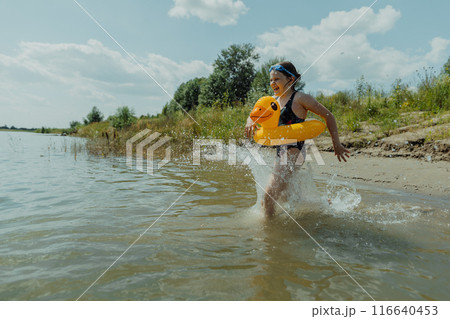 A girl is running through the water with an inflatable yellow duck on a sunny summer day. She is enjoying her time at the lake and having fun splashing around 116640453