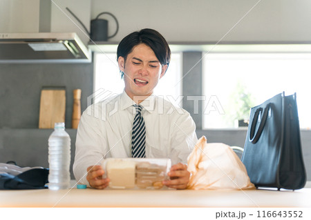 帰宅後ダイニングでコンビニ弁当を開けるスーツ姿のアジア人ビジネスマン男性 116643552