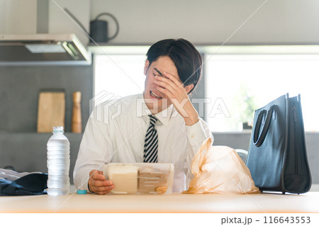 帰宅後ダイニングでコンビニ弁当を開けるスーツ姿のアジア人ビジネスマン男性 116643553
