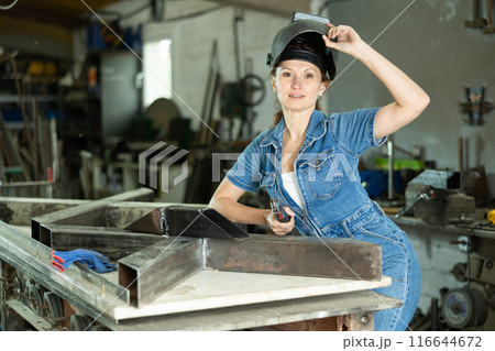 Portrait of a female welder standing with a welding semi-automatic machine and a safety helmet in metal machining workshop 116644672