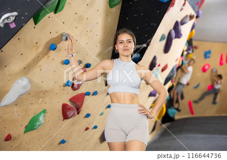 Positive slender girl in sportswear is standing near climbing wall in gym 116644736