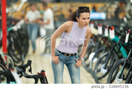 Portrait of positive woman standing in bicycle store and choosing new comfortable bike Portrait of positive woman standing in bicycle store and choosing new comfortable bike 116645136