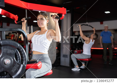 Girl doing strength training in gym, exercising upper body muscles on shoulder press machine Girl doing strength training in gym, exercising upper body muscles on shoulder press machine 116645277