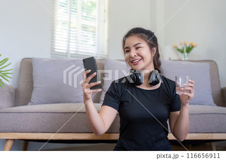 Young Woman Enjoying a Break During Home Workout with Smartphone and Water in Cozy Living Room Young Woman Enjoying a Break During Home Workout with Smartphone and Water in Cozy Living Room 116656911