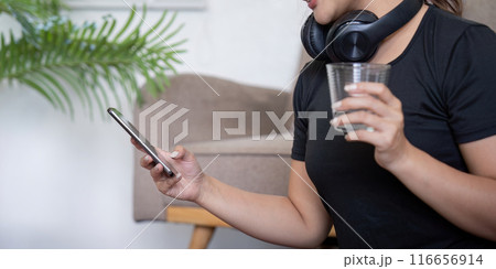 Woman with Headphones Holding a Glass of Water and Using Smartphone at Home 116656914