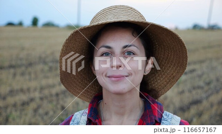 Happy smiling female farmer looks into camera standing at wheat field. Portrait of young beautiful agronomist in straw hat with barley meadow at background. Agricultural business concept Happy smiling female farmer looks into camera standing at wheat field. Portrait of young beautiful agronomist in straw hat with barley meadow at background. Agricultural business concept 116657167