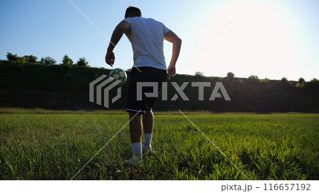 Sportsman juggling soccer ball on stadium at sunny day. Young man kicking ball at green field. Professional footballer practicing tricks at meadow with sunlight at background. Freestyle football. 116657192