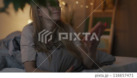 Joyful young lady taking selfie with smartphone camera posing lying in bed in bedroom in evening. Photography and modern lifestyle concept. 116657471