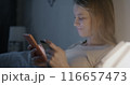 Happy young woman using smartphone and holding credit card paying in online store shopping sitting in bed in apartment in evening. E-commerce and money concept. 116657473