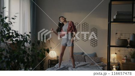 Young Asian woman dancing on bed wearing pajamas listening to music enjoying free time in bedroom in morning. Youth and entertainment concept. 116657478