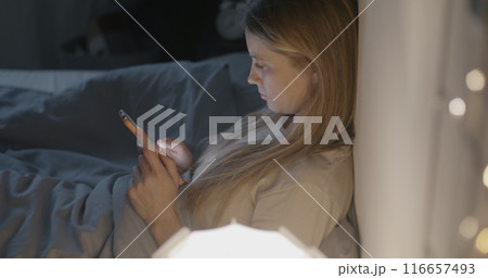 Young lady using smartphone messaging sitting in bed in dark bedroom at sleepless night. Modern technology and online communication concept. Young lady using smartphone messaging sitting in bed in dark bedroom at sleepless night. Modern technology and online communication concept. 116657493