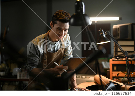 Smiling asian male artisan wearing an apron using a digital tablet in his workshop. Small business and technology concept 116657634
