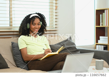 Pleased young Afro woman wearing headphone writing her diary on couch 116657662