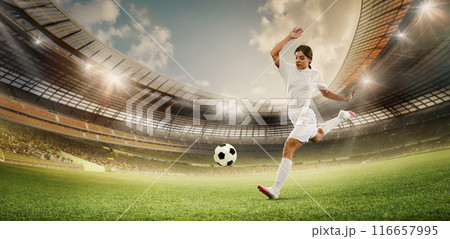 Female soccer player in white kicks ball on large stadium field. Stadium filled with spectators, with bright lights illuminating the scene. 116657995