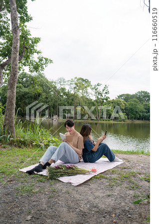 Young Couple Relaxing by a Serene Lake, Reading and Enjoying Nature on a Picnic Blanket in a Tranquil Park Setting Young Couple Relaxing by a Serene Lake, Reading and Enjoying Nature on a Picnic Blanket in a Tranquil Park Setting 116660519