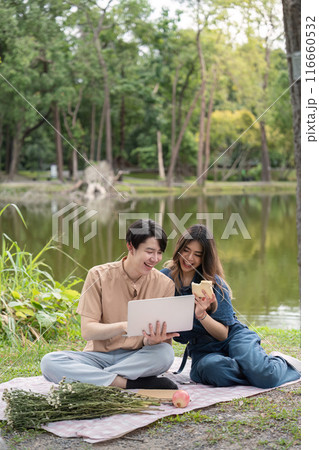 Happy Couple Enjoying a Picnic by the Lake with a Laptop and Snacks in a Serene Park Setting 116660532
