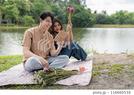 Young Couple Enjoying a Relaxing Picnic by the Lake with Flowers and Tablet on a Beautiful Day 116660538