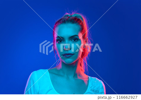 Portrait of young blonde woman looking at camera with calmness in neon light against gradient studio background. Trends. 116662927