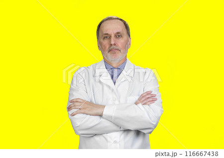 Portrait of handsome aged senior male doctor with folded arms. Isolated on yellow. 116667438