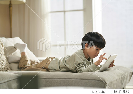little asian boy lying on front on couch playing computer game using digital tablet little asian boy lying on front on couch playing computer game using digital tablet 116671987