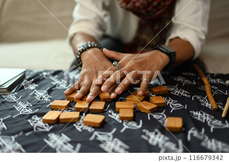 Hands of fortune teller arranging wooden runes on a table with a mystical cloth. Forecasting and divination concept Hands of fortune teller arranging wooden runes on a table with a mystical cloth. Forecasting and divination concept 116679342