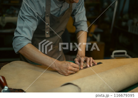 Male artisan is meticulously measuring and cutting a large piece of leather on workbench Male artisan is meticulously measuring and cutting a large piece of leather on workbench 116679395