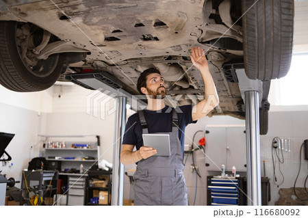 Mechanic inspecting car undercarriage on hydraulic lift in garage 116680092