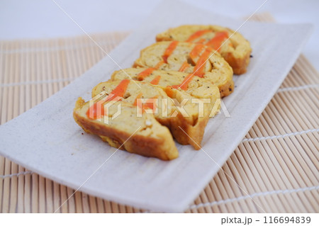 A white rectangular plate features neatly arranged slices of tamagoyaki, a Japanese rolled omelette. The omelette is drizzled with a bright orange sauce A white rectangular plate features neatly arranged slices of tamagoyaki, a Japanese rolled omelette. The omelette is drizzled with a bright orange sauce 116694839