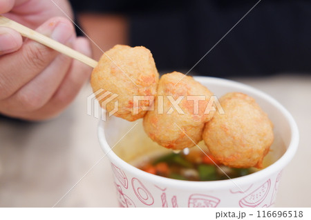 A person holds a skewer with three fried balls over a cup filled with broth and green vegetables 116696518
