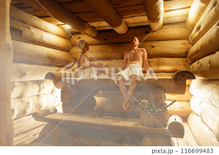Young couple relaxing and sweating in hot sauna wrapped in towel. Woman and man In Sauna. Interior of Finnish sauna, classic wooden steam room with hot steam. Russian bathroom. Relax in hot Bathhouse 116699483