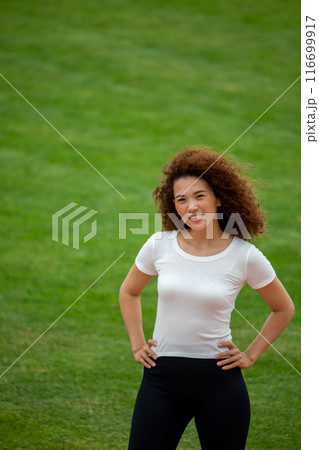 A beautiful curly girl posing in a white T-shirt and black leggings. Against the background of the stadium. Suitable for mockup 116699917
