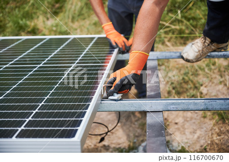 Close up of male workers in gloves installing solar panel on metal rails. Men hands mounting solar module for generating electricity through photovoltaic effect. Concept of renewable energy sources. 116700670