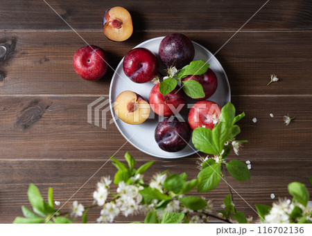 a few juicy red plums in blue plate with branch blossom flowers on a napkin on a wooden background. a few juicy red plums in blue plate with branch blossom flowers on a napkin on a wooden background. 116702136