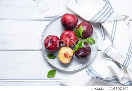 a few juicy and sweet  red plums in plate  with green leaves and napkin on a white  wood background. 116702162
