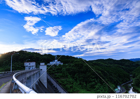 2024年夏布目ダムの夕景ダム堰堤の向こう側の山並みで最後の光を放つ夕日と上空で渦をまくようろこ雲③ 116702702