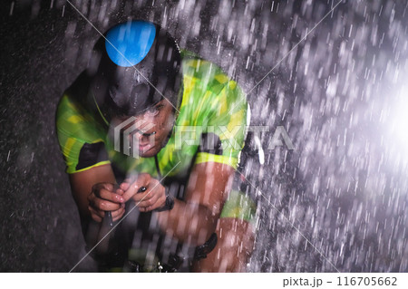 A triathlete braving the rain as he cycles through the night, preparing himself for the upcoming marathon. The blurred raindrops in the foreground and the dark, moody atmosphere in the background add 116705662