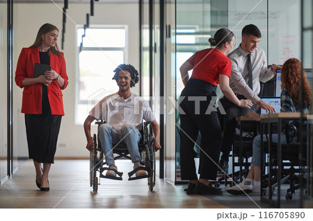 A group of young business people in a modern glass-walled office captures the essence of diversity and collaboration, while two colleagues, including an African American businessman in a wheelchair 116705890