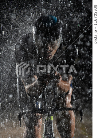 A triathlete braving the rain as he cycles through the night, preparing himself for the upcoming marathon. The blurred raindrops in the foreground and the dark, moody atmosphere in the background add 116705939