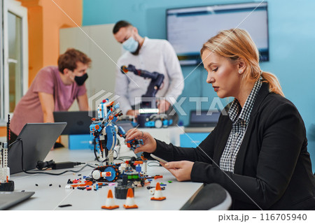 A woman sitting in a laboratory and solving problems and analyzing the robot's verification. In the background, colleagues are talking at an online meeting 116705940