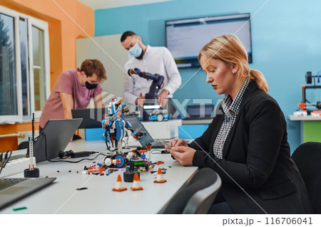 A woman sitting in a laboratory and solving problems and analyzing the robot's verification. In the background, colleagues are talking at an online meeting A woman sitting in a laboratory and solving problems and analyzing the robot's verification. In the background, colleagues are talking at an online meeting 116706041