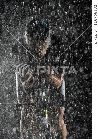 A triathlete braving the rain as he cycles through the night, preparing himself for the upcoming marathon. The blurred raindrops in the foreground and the dark, moody atmosphere in the background add A triathlete braving the rain as he cycles through the night, preparing himself for the upcoming marathon. The blurred raindrops in the foreground and the dark, moody atmosphere in the background add 116706332
