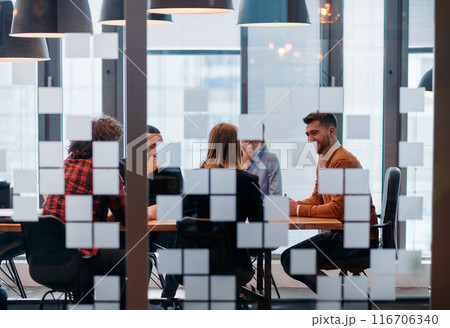In a sleek glass office space, a team of business professionals engage in lively discussions and exchange strategic ideas, showcasing collaboration and innovation at the heart of modern corporate 116706340