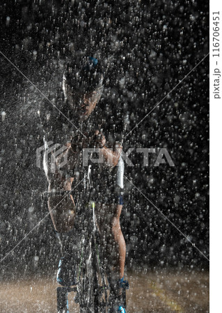A triathlete braving the rain as he cycles through the night, preparing himself for the upcoming marathon. The blurred raindrops in the foreground and the dark, moody atmosphere in the background add 116706451