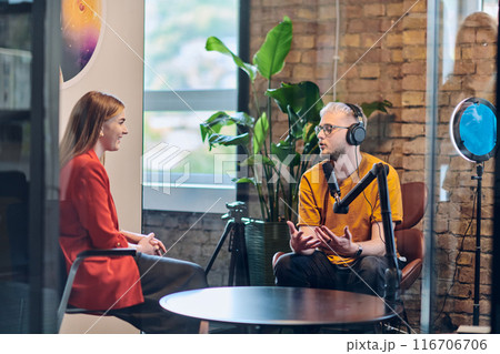 A gathering of young business professionals, some seated in a glass-walled office, engage in a lively conversation and record an online podcast, embodying modern collaboration and dynamic interaction 116706706
