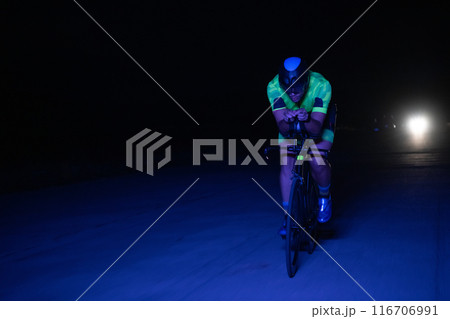 A triathlete rides his bike in the darkness of night, pushing himself to prepare for a marathon. The contrast between the darkness and the light of his bike creates a sense of drama and highlights the A triathlete rides his bike in the darkness of night, pushing himself to prepare for a marathon. The contrast between the darkness and the light of his bike creates a sense of drama and highlights the 116706991