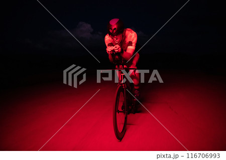 A triathlete rides his bike in the darkness of night, pushing himself to prepare for a marathon. The contrast between the darkness and the light of his bike creates a sense of drama and highlights the 116706993