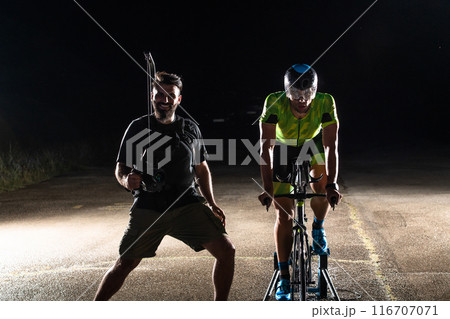 Behind the scenes. Professional videographer on a night shoot with a triathlete riding a bicycle for video recording purposes Behind the scenes. Professional videographer on a night shoot with a triathlete riding a bicycle for video recording purposes 116707071