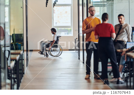 A diverse group of colleagues engages in a discussion about business challenges within a modern coworking startup center, while in the background, their wheelchair-bound colleague symbolizes 116707643