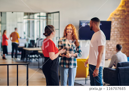 A group of young business individuals, including a girl with orange hair and an African American man, stands in a modern corporate hallway, collectively examining business progress on a smartphone 116707685
