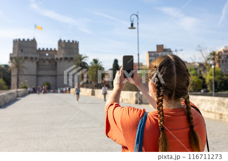 Redhead female traveler in casual dress taking picture of Torres de Serranos tower with smartphone, travel in Europe. Attractive female tourist is exploring new city. Hight quality photo, rear view 116711273